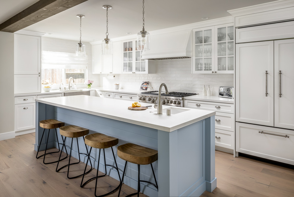 Photo of a large beach style l-shaped kitchen in Sacramento with glass-front cabinets, white splashback, with island, a single-bowl sink, white cabinets, panelled appliances, light hardwood floors and beige floor.