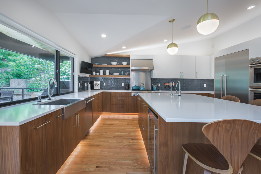This is an example of a midcentury u-shaped eat-in kitchen in Seattle with a farmhouse sink, flat-panel cabinets, dark wood cabinets, quartz benchtops, blue splashback, subway tile splashback, stainless steel appliances, medium hardwood floors, with island, white benchtop and vaulted.