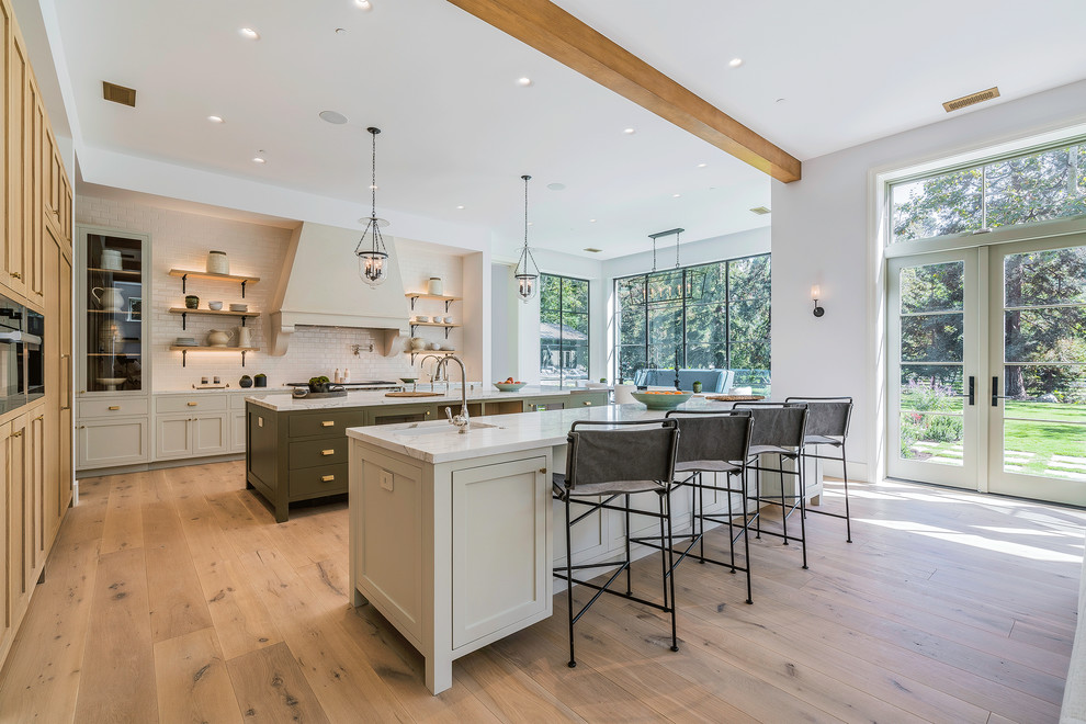 Photo of a large country single-wall eat-in kitchen in Los Angeles with a farmhouse sink, recessed-panel cabinets, marble benchtops, white splashback, subway tile splashback, stainless steel appliances, light hardwood floors, multiple islands, beige floor and light wood cabinets.