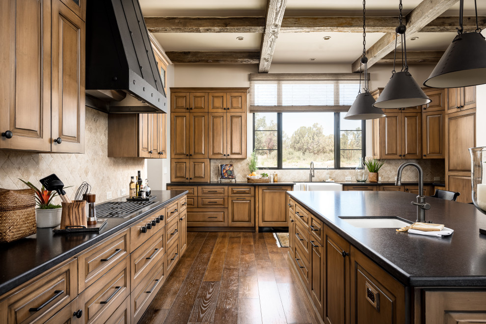 Southwest u-shaped dark wood floor, brown floor and exposed beam kitchen photo in Phoenix with a farmhouse sink, raised-panel cabinets, medium tone wood cabinets, beige backsplash, paneled appliances, an island and black countertops
