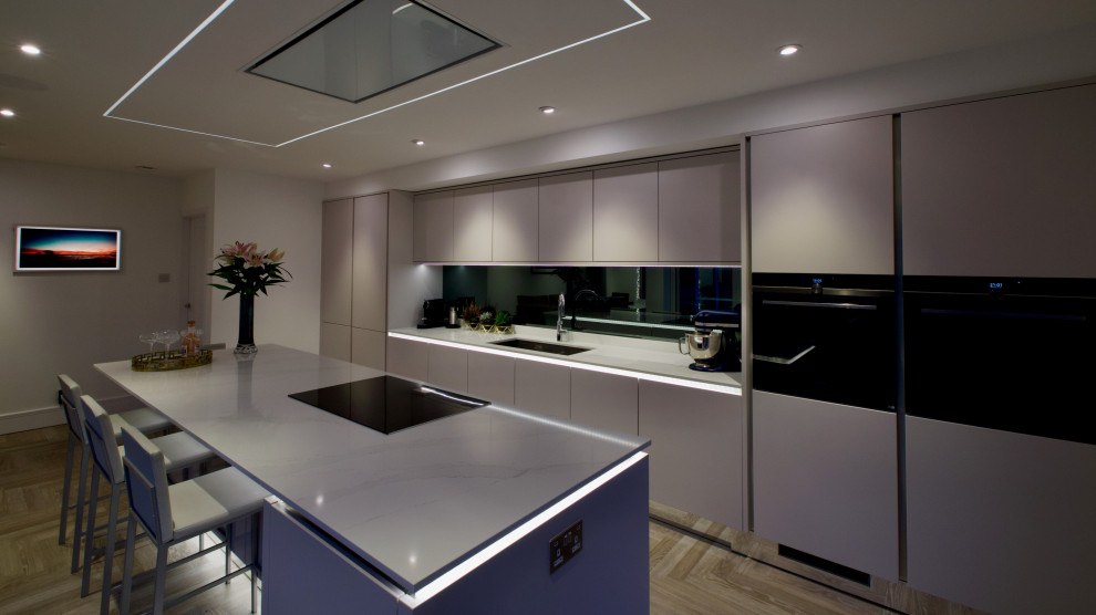 Идея дизайна: кухня-гостиная: освещение в стиле модернизм с врезной мойкой, плоскими фасадами, серым фартуком, зеркальным фартуком, черной техникой, островом и белой столешницей