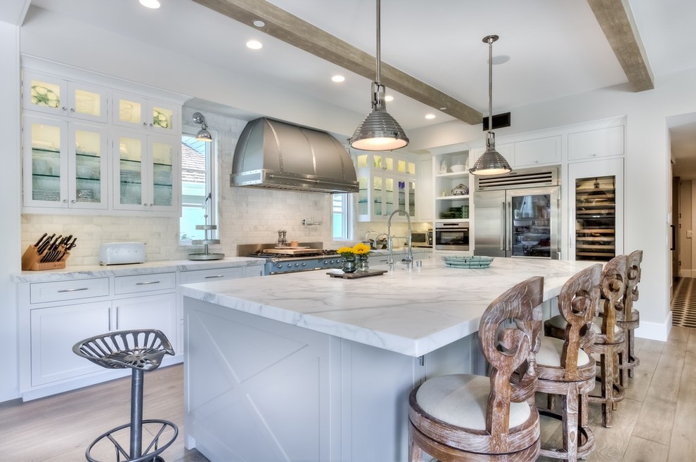 This is an example of a large country l-shaped open plan kitchen in Orange County with recessed-panel cabinets, white cabinets, marble benchtops, white splashback, subway tile splashback, stainless steel appliances, with island, a farmhouse sink and light hardwood floors.