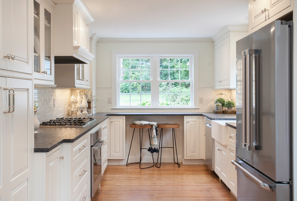 This is an example of a mid-sized country u-shaped separate kitchen in Bridgeport with raised-panel cabinets, white cabinets, no island, black benchtop, a farmhouse sink, granite benchtops, white splashback, subway tile splashback, stainless steel appliances and light hardwood floors.