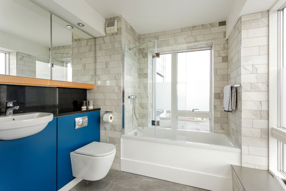 Mid-sized contemporary master bathroom in Dublin with flat-panel cabinets, light wood cabinets, a drop-in tub, a wall-mount toilet, gray tile, porcelain tile, grey walls, porcelain floors, an integrated sink, granite benchtops, grey floor and black benchtops.