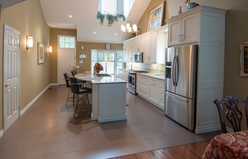 Eat-in kitchen - mid-sized transitional galley ceramic tile eat-in kitchen idea in Toronto with a double-bowl sink, shaker cabinets, white cabinets, beige backsplash, stone tile backsplash, stainless steel appliances, an island and granite countertops