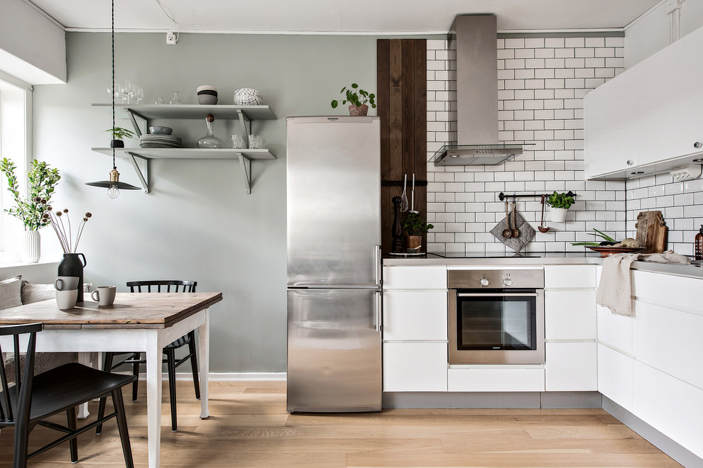 Design ideas for a mid-sized scandinavian l-shaped eat-in kitchen in Gothenburg with flat-panel cabinets, white cabinets, white splashback, subway tile splashback, stainless steel appliances, light hardwood floors, no island and beige floor.