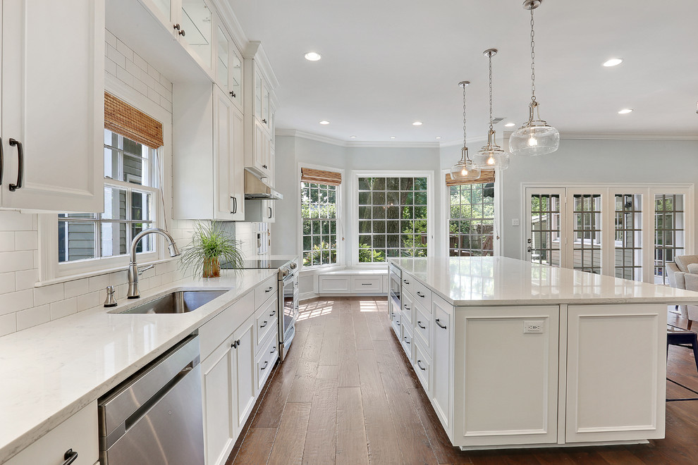 This is an example of a mid-sized transitional l-shaped open plan kitchen in New Orleans with an undermount sink, white cabinets, quartz benchtops, white splashback, subway tile splashback, stainless steel appliances, with island, brown floor, white benchtop, recessed-panel cabinets and medium hardwood floors.