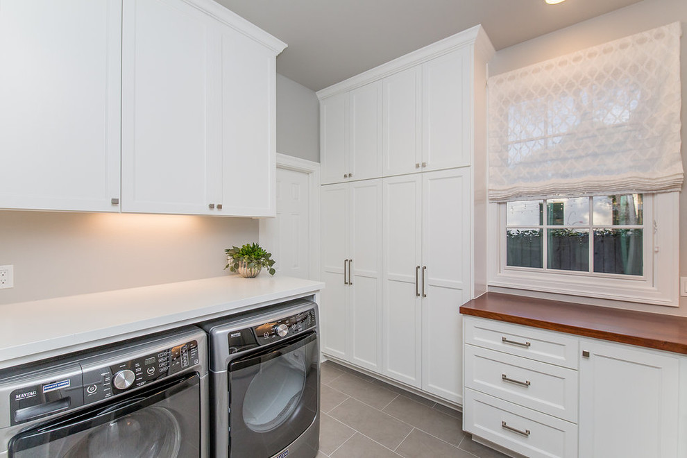 Inspiration for a large transitional l-shaped utility room in Austin with shaker cabinets, white cabinets, wood benchtops, grey walls, a side-by-side washer and dryer, grey floor, porcelain floors and brown benchtop.