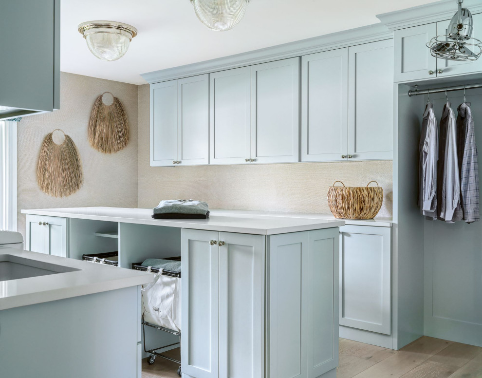 Inspiration for a large transitional galley dedicated laundry room in St Louis with an undermount sink, flat-panel cabinets, blue cabinets, quartz benchtops, beige walls, light hardwood floors, a side-by-side washer and dryer, brown floor, white benchtop and wallpaper.