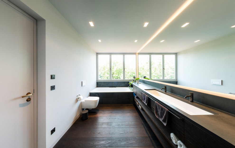 This is an example of an expansive modern 3/4 bathroom in Munich with flat-panel cabinets, a drop-in tub, a wall-mount toilet, grey walls, medium hardwood floors, solid surface benchtops, brown floor, dark wood cabinets, black tile, an integrated sink and brown benchtops.