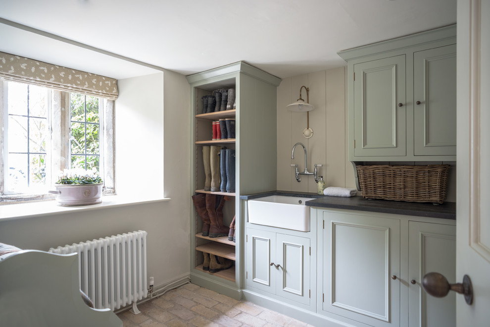 Large country galley laundry room in Dorset with a farmhouse sink, recessed-panel cabinets, grey cabinets, granite benchtops, beige walls, brick floors, beige floor and black benchtop.