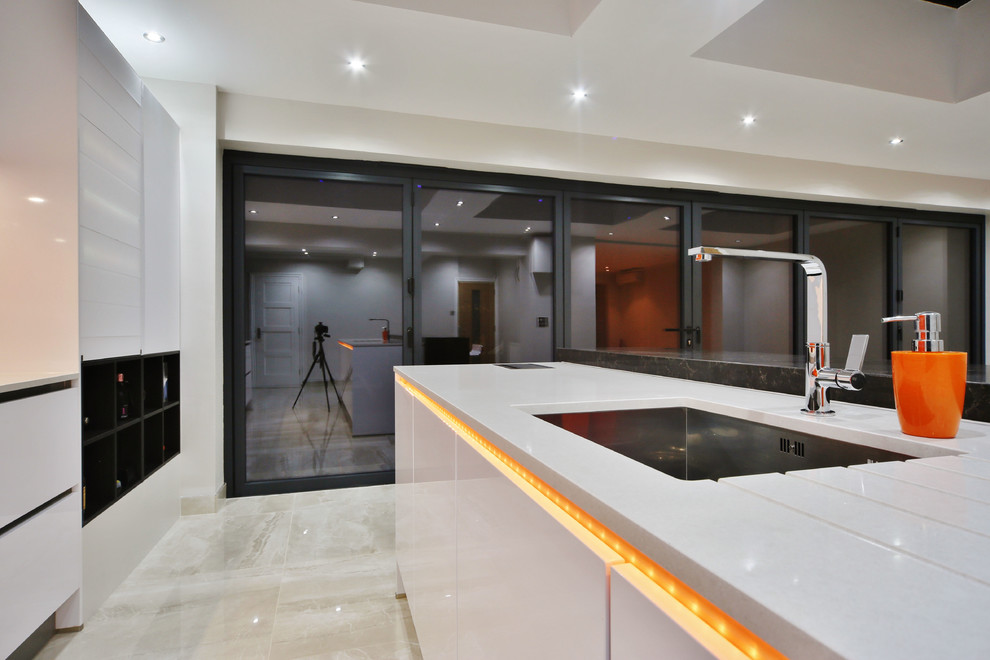 Design ideas for a mid-sized contemporary single-wall open plan kitchen in London with an undermount sink, flat-panel cabinets, white cabinets, quartzite benchtops, orange splashback, glass sheet splashback, black appliances, porcelain floors, with island, beige floor and white benchtop.