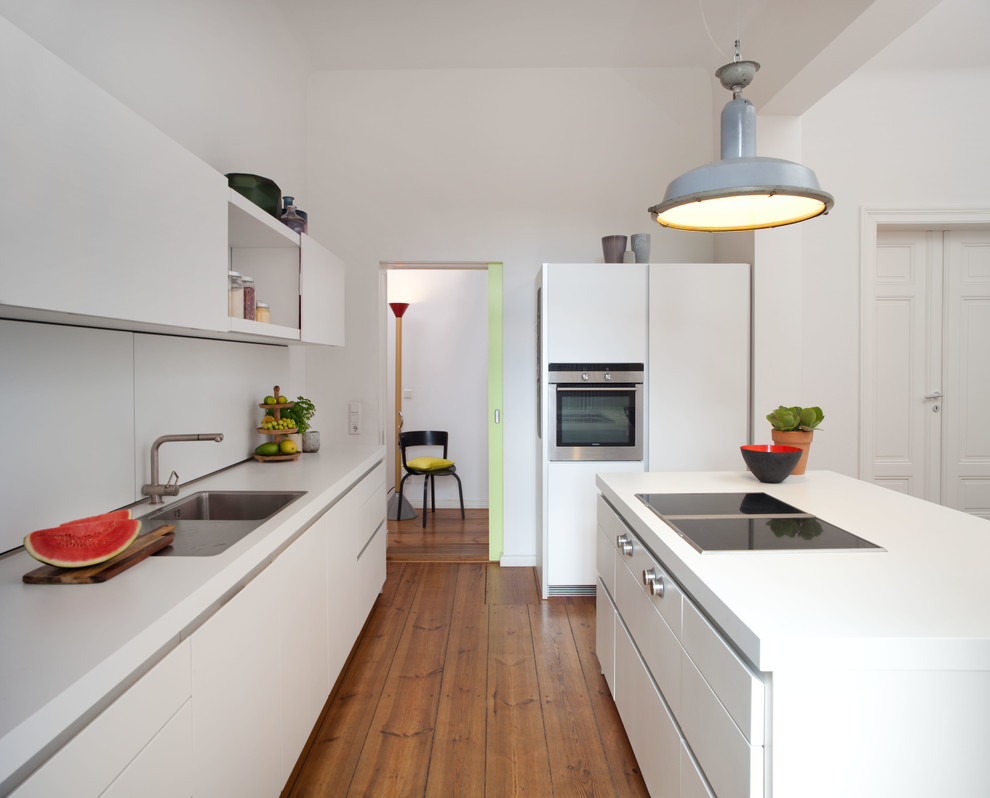 Grifflose Küche mit Einbauwaschbecken, flächenbündigen Schrankfronten, weißen Schränken, Mineralwerkstoff-Arbeitsplatte, Elektrogeräten mit Frontblende, braunem Holzboden und Kücheninsel in Berlin