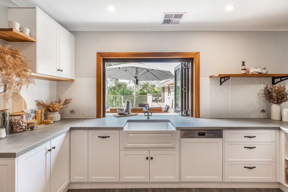 Large country u-shaped kitchen pantry in Adelaide with a farmhouse sink, shaker cabinets, white cabinets, quartz benchtops, white splashback, metal splashback, black appliances, ceramic floors, a peninsula, grey floor and grey benchtop.