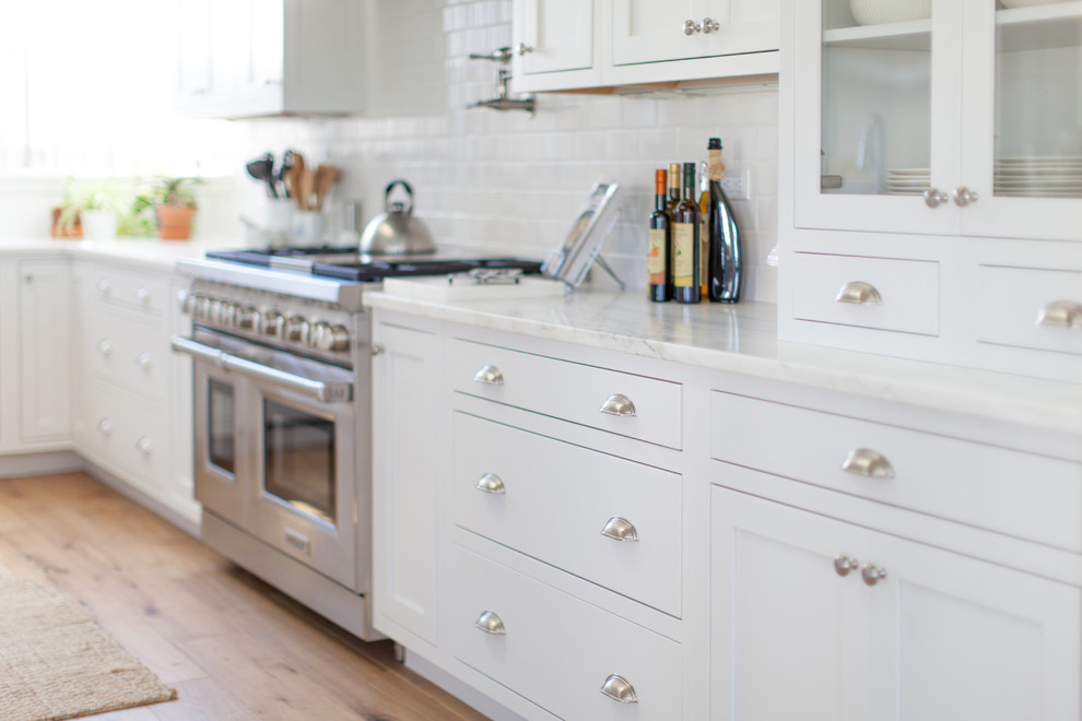 Example of a large transitional l-shaped light wood floor eat-in kitchen design in Phoenix with a farmhouse sink, white cabinets, white backsplash, stainless steel appliances, an island, shaker cabinets, stainless steel countertops and porcelain backsplash