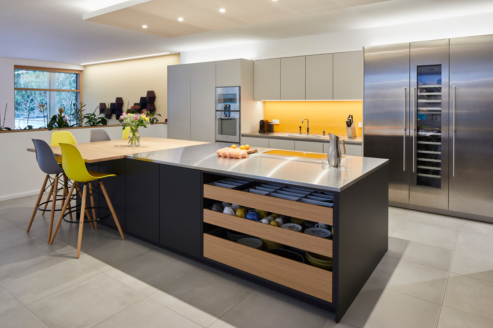 Inspiration for a large modern single-wall open plan kitchen with black cabinets, stainless steel benchtops, yellow splashback, glass sheet splashback, stainless steel appliances, ceramic floors, with island and grey floor.