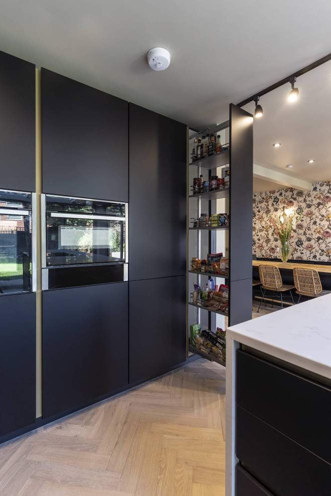 This is an example of a small scandinavian galley open plan kitchen in Oxfordshire with a submerged sink, beaded cabinets, black cabinets, quartz worktops, white splashback, engineered quartz splashback, black appliances, an island, brown floors, white worktops, a vaulted ceiling and feature lighting.