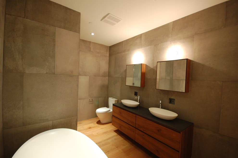 Photo of a mid-sized modern master bathroom in New York with flat-panel cabinets, dark wood cabinets, a freestanding tub, a one-piece toilet, gray tile, cement tile, grey walls, medium hardwood floors, a vessel sink, soapstone benchtops, brown floor and black benchtops.