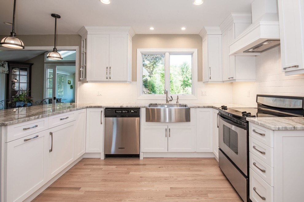 This is an example of a large transitional u-shaped eat-in kitchen in New York with a farmhouse sink, shaker cabinets, white cabinets, marble benchtops, white splashback, subway tile splashback, stainless steel appliances, light hardwood floors, a peninsula, beige floor and grey benchtop.