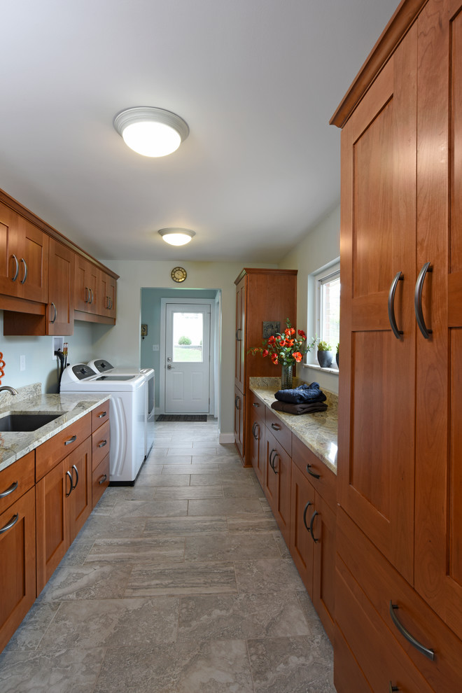 Photo of a large traditional galley utility room in Other with an undermount sink, shaker cabinets, medium wood cabinets, granite benchtops, grey walls, porcelain floors, a side-by-side washer and dryer and brown floor.