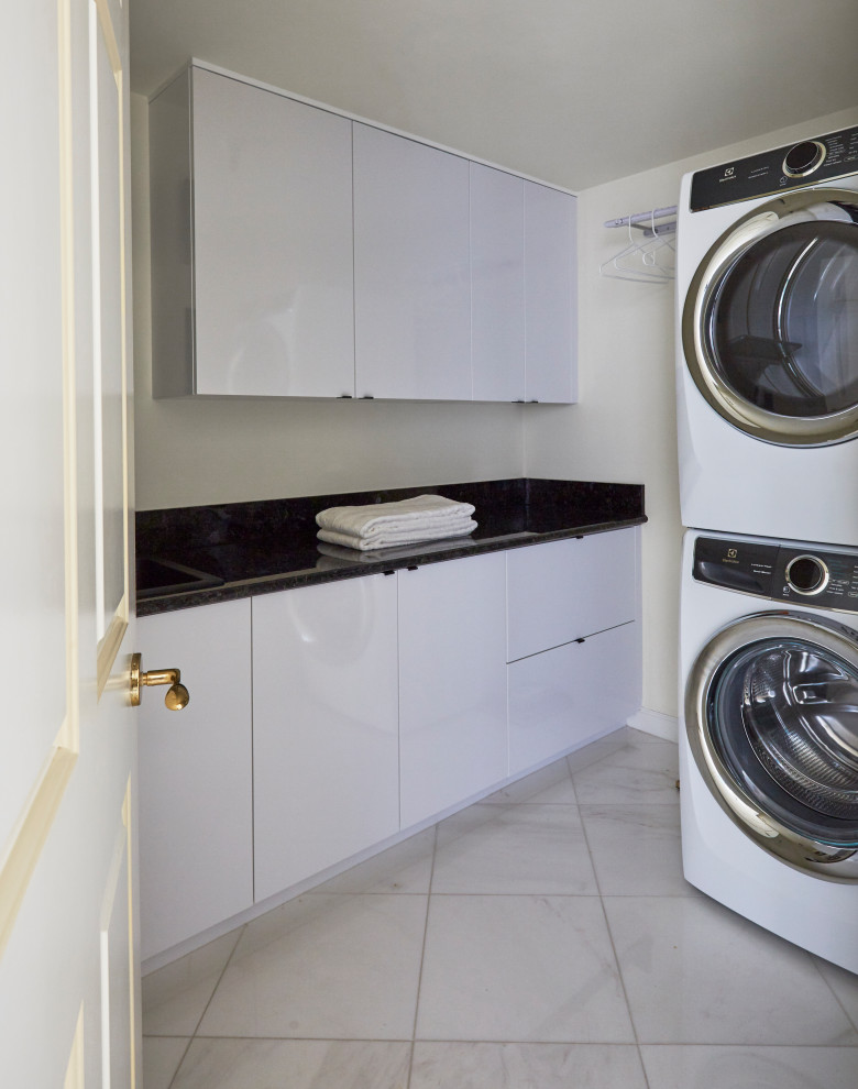 Small contemporary single-wall dedicated laundry room in Miami with an undermount sink, flat-panel cabinets, white cabinets, granite benchtops, black splashback, granite splashback, white walls, marble floors, a stacked washer and dryer, white floor and black benchtop.
