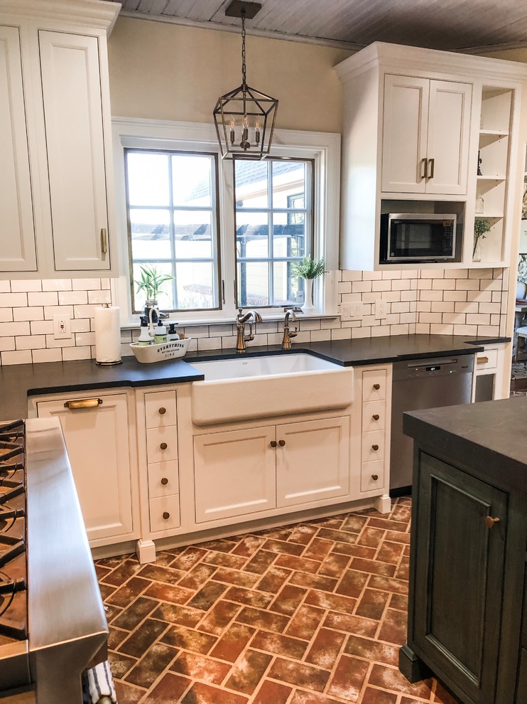 Mid-sized country u-shaped eat-in kitchen in Philadelphia with a farmhouse sink, recessed-panel cabinets, white cabinets, soapstone benchtops, white splashback, subway tile splashback, stainless steel appliances, brick floors, with island, red floor and black benchtop.