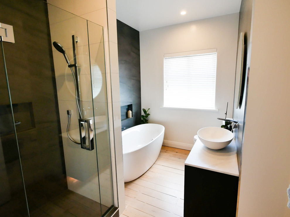 Inspiration for a mid-sized contemporary master bathroom in Auckland with recessed-panel cabinets, black cabinets, a freestanding tub, a corner shower, a two-piece toilet, gray tile, porcelain tile, white walls, light hardwood floors, a vessel sink, limestone benchtops, brown floor, a hinged shower door and white benchtops.