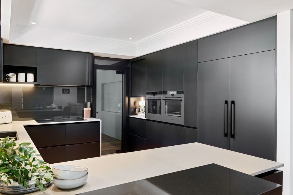 This is an example of a large contemporary u-shaped open plan kitchen in Perth with a submerged sink, flat-panel cabinets, black cabinets, engineered stone countertops, black splashback, glass sheet splashback, black appliances, light hardwood flooring, an island, brown floors, grey worktops and a drop ceiling.