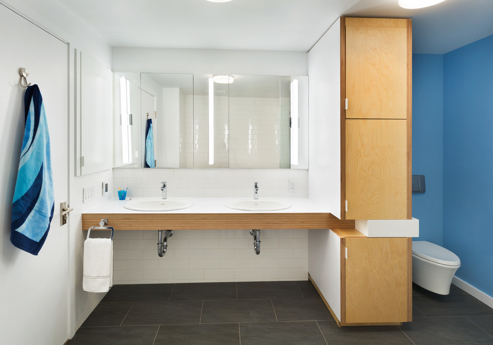 This is an example of a mid-sized contemporary kids bathroom in New York with a drop-in sink, flat-panel cabinets, laminate benchtops, a wall-mount toilet, white tile, stone tile, blue walls, slate floors and medium wood cabinets.