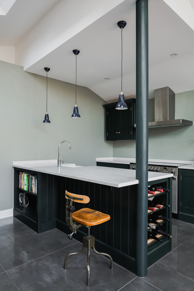 This is an example of a transitional galley kitchen in Hertfordshire with a farmhouse sink, raised-panel cabinets, black cabinets, grey splashback, stainless steel appliances, a peninsula, grey floor and white benchtop.