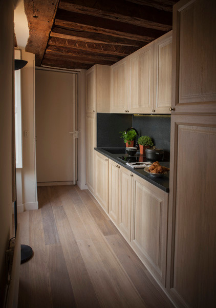 This is an example of a small transitional single-wall open plan kitchen in Paris with an integrated sink, beaded inset cabinets, light wood cabinets, granite benchtops, black splashback, limestone splashback, panelled appliances, light hardwood floors, no island, brown floor and black benchtop.