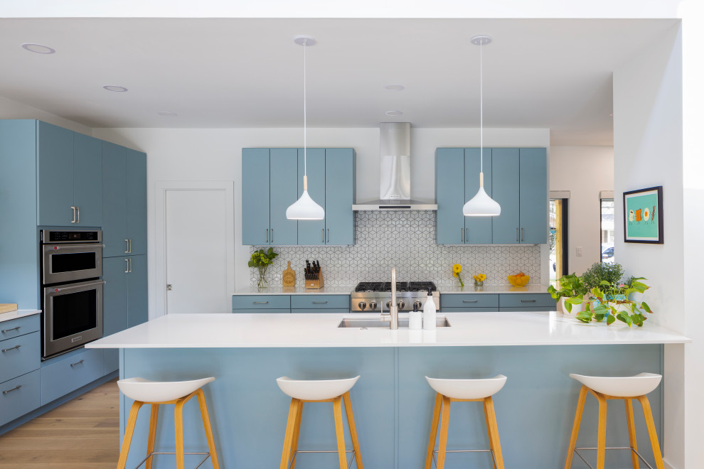 This is an example of a large scandinavian l-shaped open plan kitchen in Austin with an undermount sink, flat-panel cabinets, blue cabinets, quartz benchtops, white splashback, mosaic tile splashback, stainless steel appliances, light hardwood floors, a peninsula and white benchtop.