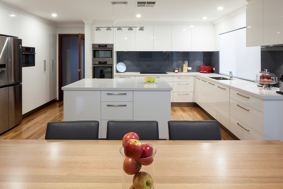 This is an example of a modern l-shaped eat-in kitchen in Perth with flat-panel cabinets, grey cabinets, quartz benchtops, glass sheet splashback, stainless steel appliances, with island, black splashback, white benchtop and light hardwood floors.