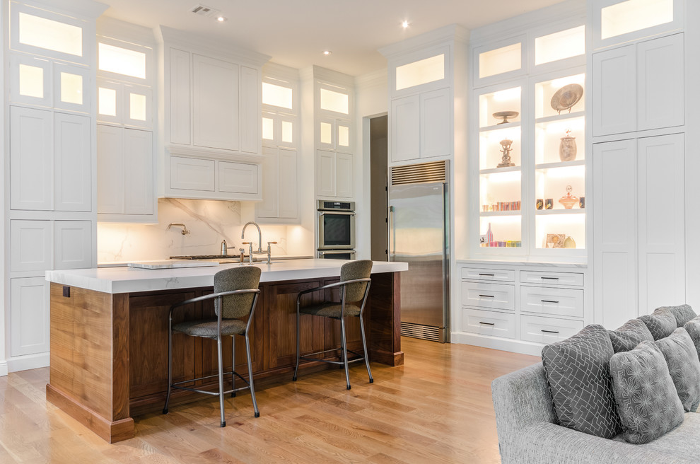 Example of a mid-sized transitional l-shaped light wood floor and beige floor kitchen design with glass-front cabinets, white cabinets, white backsplash, stainless steel appliances, an island and white countertops