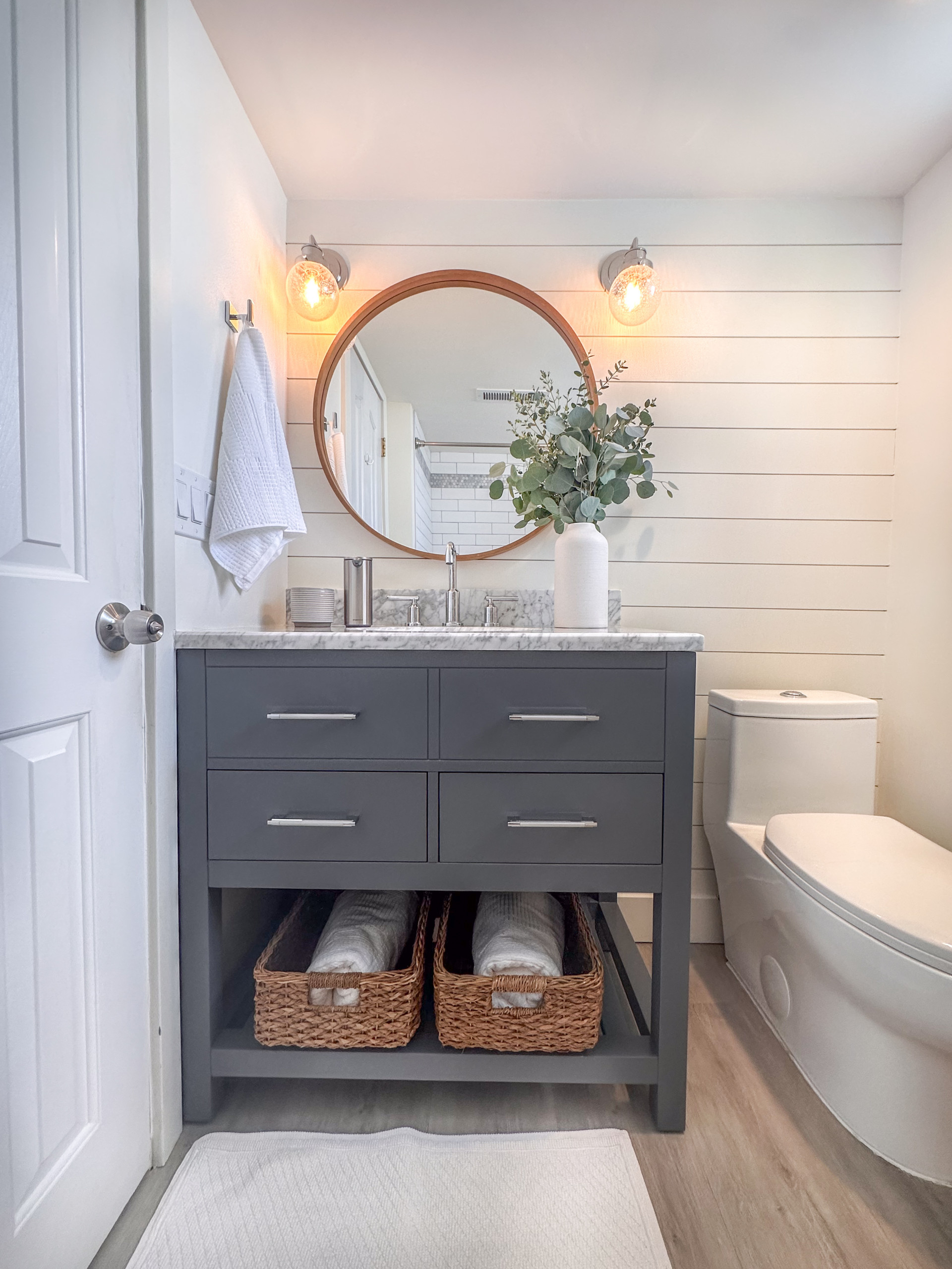 Modern farmhouse bathroom remodel featuring a beautiful Carrara marble counter and gray vanity which includes two drawers and an open shelf at the bottom for wicker baskets that add warmth and texture