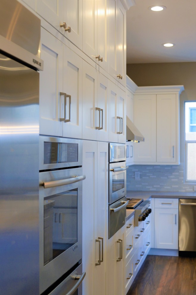 This is an example of a large transitional l-shaped open plan kitchen in Las Vegas with a farmhouse sink, recessed-panel cabinets, white cabinets, quartzite benchtops, white splashback, glass tile splashback, stainless steel appliances, medium hardwood floors and with island.