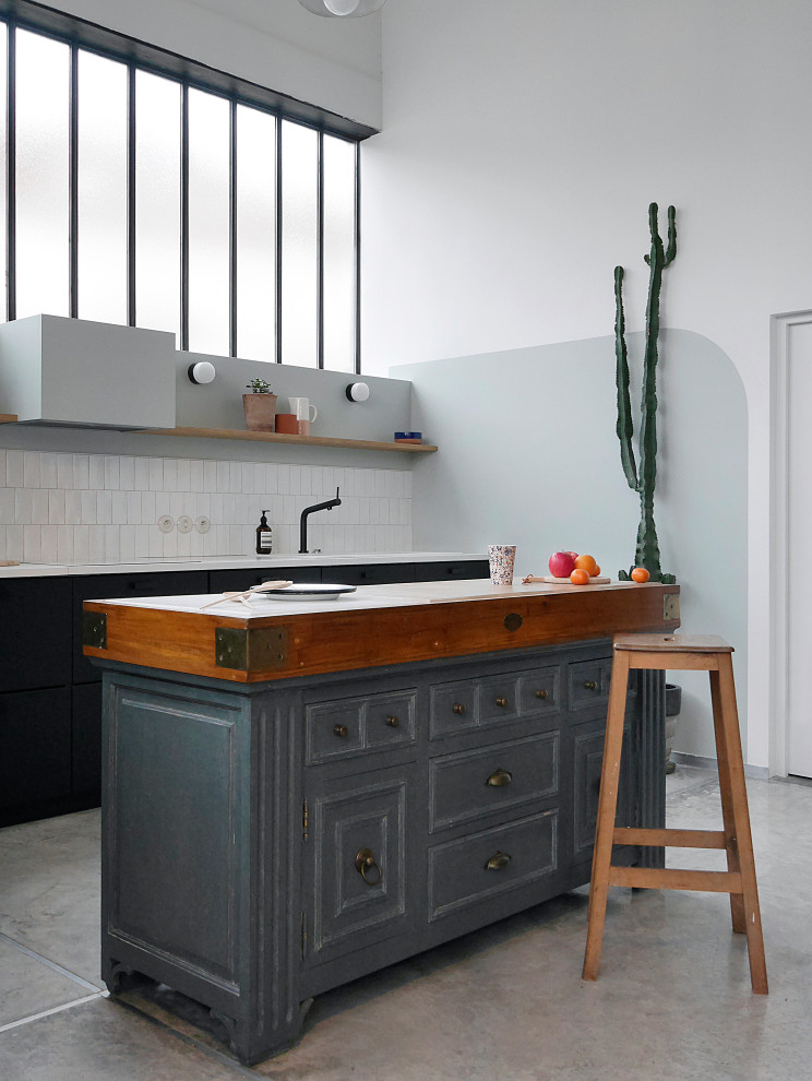 This is an example of an expansive industrial l-shaped open plan kitchen with a single-bowl sink, beaded inset cabinets, black cabinets, laminate benchtops, white splashback, ceramic splashback, black appliances, concrete floors, with island, grey floor and white benchtop.