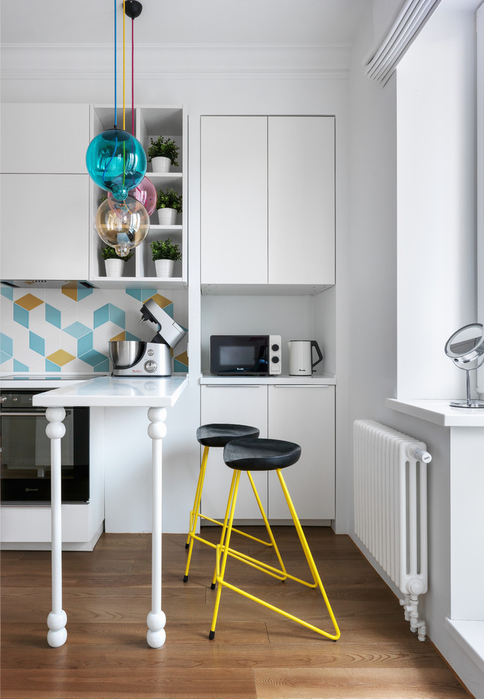 This is an example of a contemporary single-wall separate kitchen in Moscow with flat-panel cabinets, white cabinets, multi-coloured splashback, cement tile splashback, black appliances, medium hardwood floors, a peninsula, white benchtop and brown floor.