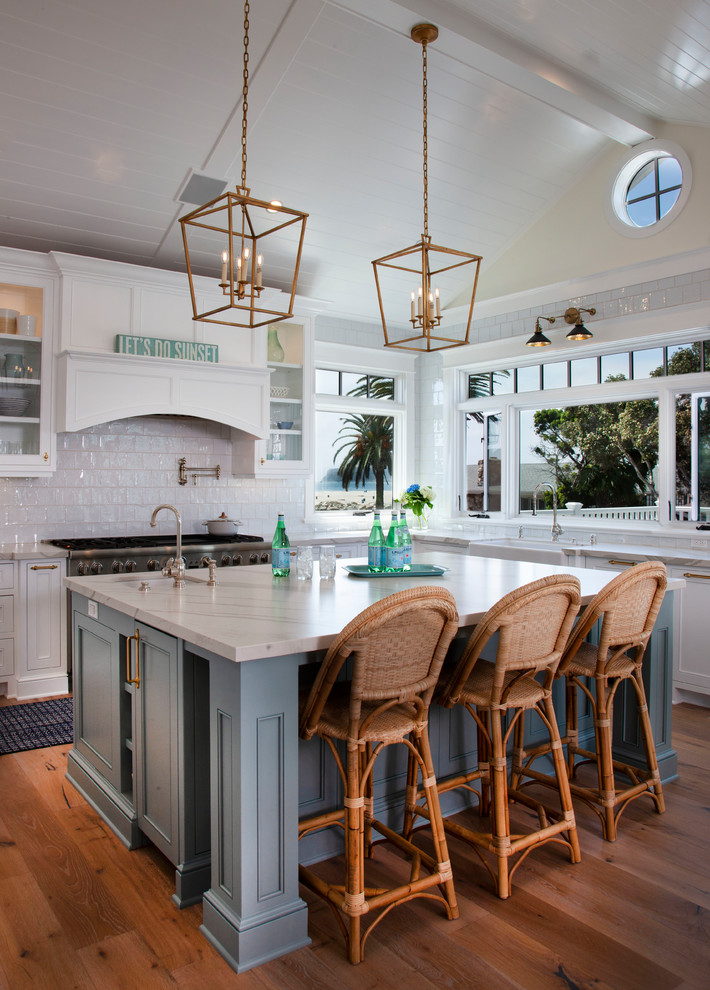 Design ideas for a mid-sized beach style l-shaped eat-in kitchen in San Diego with a farmhouse sink, shaker cabinets, quartz benchtops, white splashback, ceramic splashback, stainless steel appliances, with island, medium hardwood floors, brown floor, white benchtop and white cabinets.