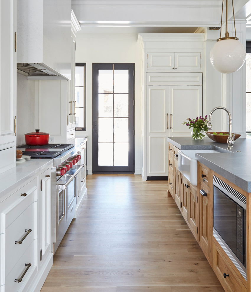 This is an example of an expansive traditional u-shaped open plan kitchen in Chicago with a farmhouse sink, recessed-panel cabinets, white cabinets, quartz benchtops, white splashback, stone slab splashback, stainless steel appliances, light hardwood floors, with island, brown floor and grey benchtop.