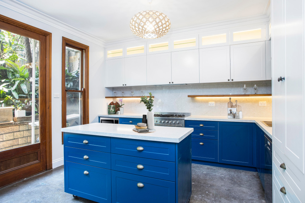 Photo of a mid-sized beach style l-shaped eat-in kitchen in Sydney with a drop-in sink, shaker cabinets, blue cabinets, quartz benchtops, white splashback, stone slab splashback, stainless steel appliances, concrete floors, with island, grey floor and white benchtop.