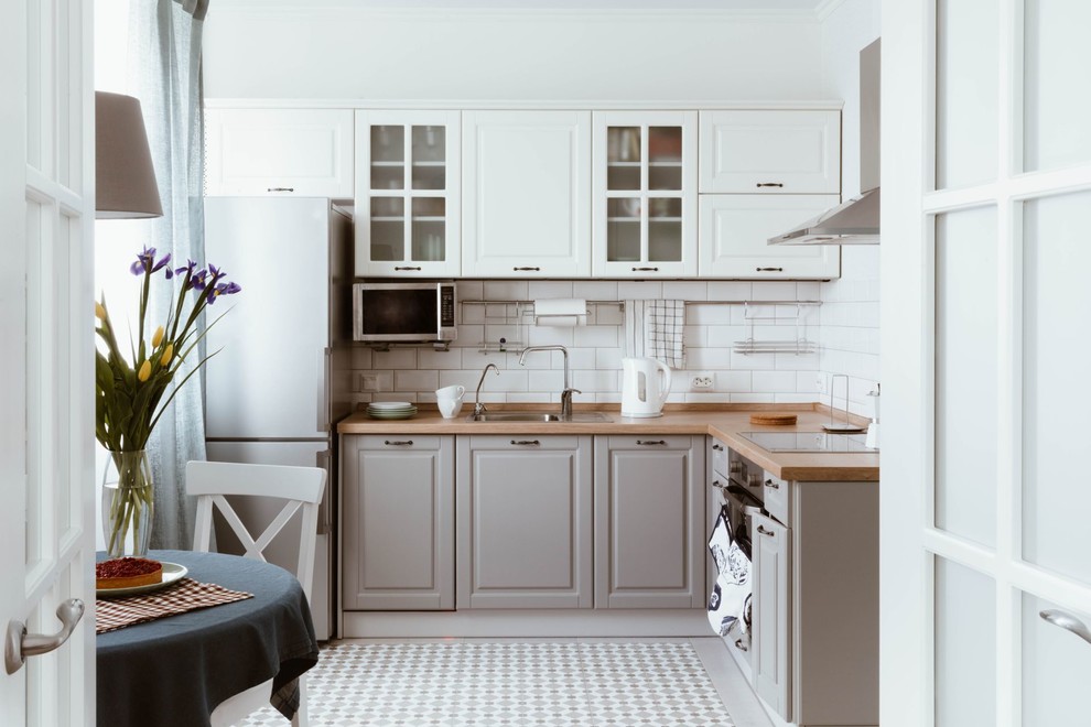 This is an example of a transitional l-shaped separate kitchen in Other with a drop-in sink, raised-panel cabinets, grey cabinets, wood benchtops, white splashback, subway tile splashback, black appliances, no island, grey floor and brown benchtop.