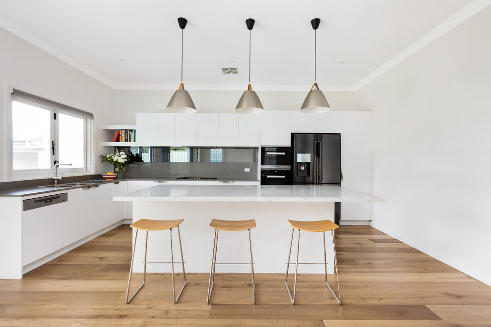 Design ideas for a contemporary l-shaped kitchen in Sydney with a drop-in sink, flat-panel cabinets, white cabinets, grey splashback, black appliances, light hardwood floors, with island and grey benchtop.