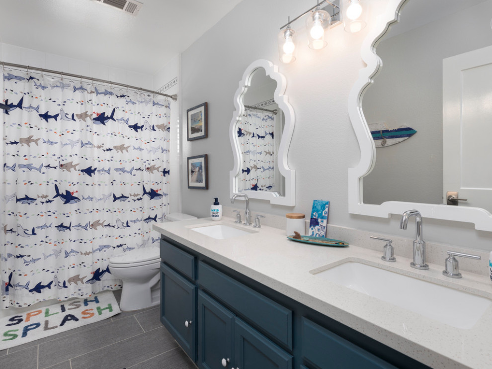 Inspiration for a mid-sized beach style kids bathroom in Los Angeles with shaker cabinets, blue cabinets, a one-piece toilet, subway tile, grey walls, porcelain floors, an undermount sink, engineered quartz benchtops, a shower curtain, white benchtops, a double vanity, a built-in vanity, a drop-in tub, a shower/bathtub combo, white tile and grey floor.
