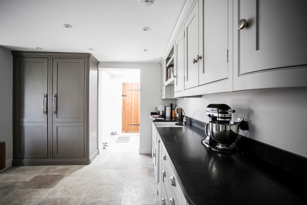Mid-sized traditional u-shaped open plan kitchen in Kent with a farmhouse sink, shaker cabinets, grey cabinets, granite benchtops, brown splashback, ceramic splashback, black appliances, limestone floors, with island, beige floor and black benchtop.