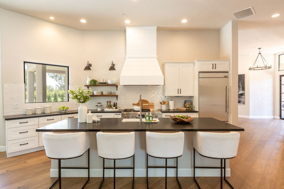 Example of a transitional l-shaped medium tone wood floor and brown floor kitchen design in Phoenix with shaker cabinets, white cabinets, white backsplash, stainless steel appliances, an island and black countertops