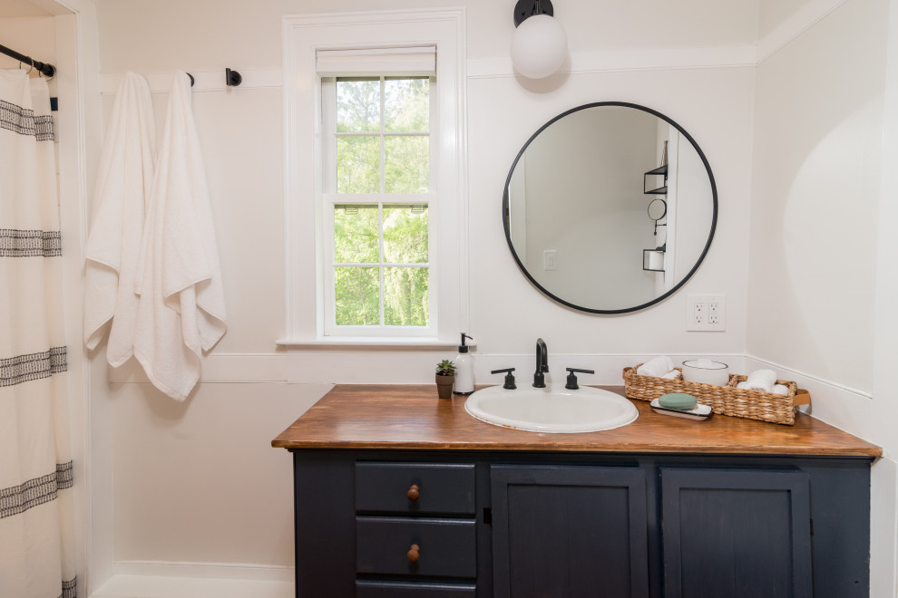 Photo of a mid-sized country master bathroom in New York with shaker cabinets, blue cabinets, an alcove tub, an alcove shower, a two-piece toilet, white walls, medium hardwood floors, a drop-in sink, wood benchtops, a shower curtain, brown benchtops, a single vanity and a built-in vanity.