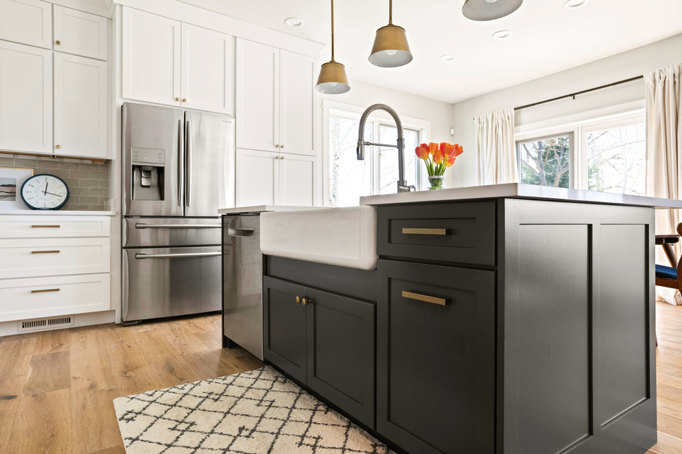 Transitional l-shaped light wood floor and brown floor kitchen photo with a farmhouse sink, shaker cabinets, white cabinets, quartz countertops, white backsplash, subway tile backsplash, stainless steel appliances, an island and white countertops
