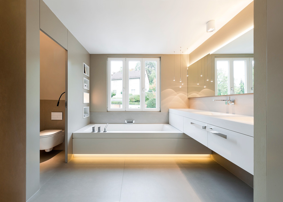 This is an example of a large contemporary bathroom in Stuttgart with an integrated sink, flat-panel cabinets, white cabinets, a wall-mount toilet, beige walls, solid surface benchtops and an alcove tub.