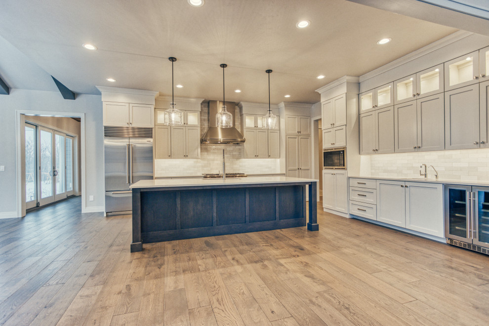 Example of a transitional l-shaped medium tone wood floor and brown floor kitchen pantry design with an undermount sink, shaker cabinets, white cabinets, quartz countertops, white backsplash, ceramic backsplash, stainless steel appliances, an island and white countertops